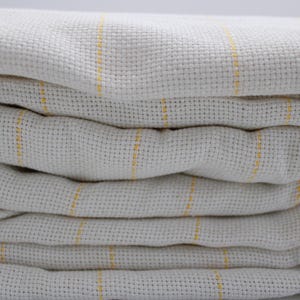 May include: A stack of folded, off-white fabric with a woven texture. Thin, vertical yellow stripes run across the fabric, adding a subtle color contrast. The fabric appears soft and slightly rumpled.