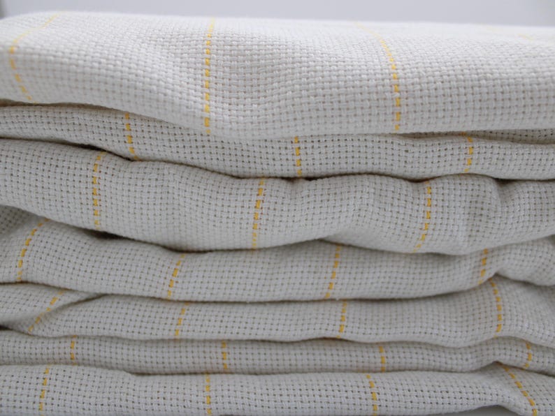 May include: A stack of folded, off-white fabric with a woven texture. Thin, vertical yellow stripes run across the fabric, adding a subtle color contrast. The fabric appears soft and slightly rumpled.