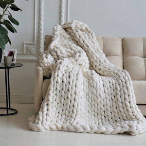 May include: A chunky knit ivory blanket draped over a beige sofa. The blanket's thick, oversized stitches create a textured, cozy appearance. The blanket is made of a soft, wool-like material, perfect for adding warmth and style to any living space.