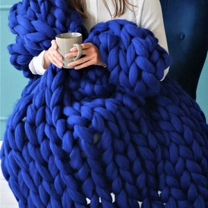 May include: A large, chunky-knit blanket in a vibrant royal blue. The blanket is draped over a person seated in a blue chair, with a gray mug in their hands. The thick yarn creates a cozy, textured appearance, perfect for adding warmth and style to any space.