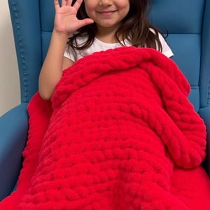 Girl on blue sofa waving hand and covered in red chenille blanket