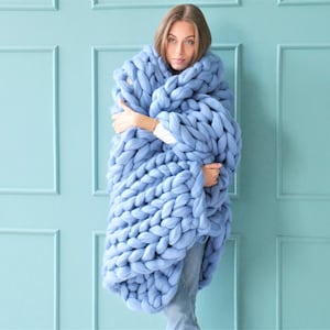 May include: A large, chunky-knit blanket in a soft, light blue color. The blanket is wrapped around a person, showcasing its thick, oversized weave. The blanket appears to be made of a soft, possibly wool, material, perfect for cozy comfort.