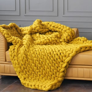 May include: A chunky knit blanket in a vibrant mustard yellow color is draped over a tan leather sofa. The blanket's large, woven texture is prominently displayed, creating a cozy and inviting aesthetic. The background features a gray wall.