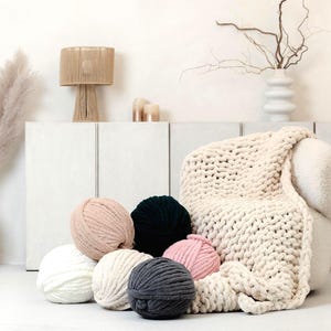 Cream chunky knit chenille blanket laying down on the sofa as a home decor detail in a modern living room together with super chunky chenille yarns for knitting and crocheting. This soft chenille blanket is hand knitted exactly from this plush chenille yarn.