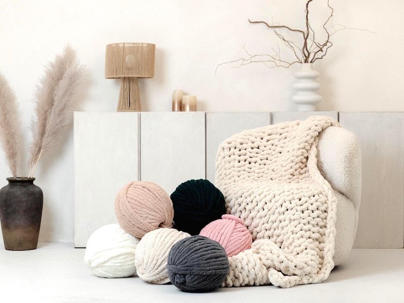 Cream chunky knit chenille blanket laying down on the sofa as a home decor detail in a modern living room together with super chunky chenille yarns for knitting and crocheting. This soft chenille blanket is hand knitted exactly from this plush chenille yarn.