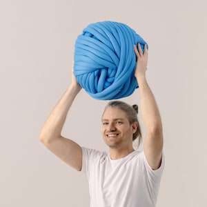 May include: A large, spherical ball of thick, bright blue yarn made from soft, tubular fabric. The yarn is wound into a dense, continuous coil, creating a smooth, rounded texture that is held up high with both hands against a plain background.