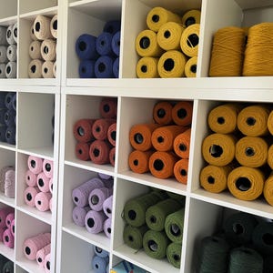 A white shelving unit filled with spools of yarn in various colors. The yarn colors include white, blue, red, orange, yellow, green, and black. The yarn is neatly organized in individual cubbies.