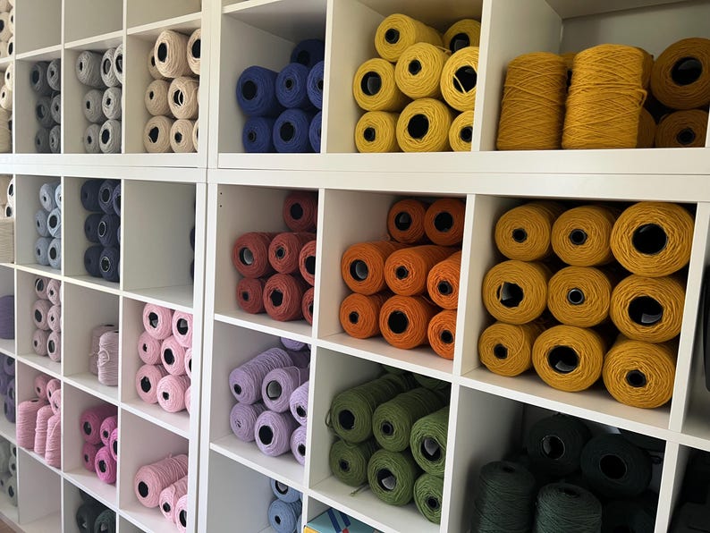 A white shelving unit filled with spools of yarn in various colors. The yarn colors include white, blue, red, orange, yellow, green, and black. The yarn is neatly organized in individual cubbies.