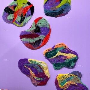 May include: Seven abstract, organic-shaped rugs in various colors, including purple, green, red, and yellow, are displayed against a lavender wall. The rugs have a textured, woven appearance, with each piece showcasing a unique color palette and design.