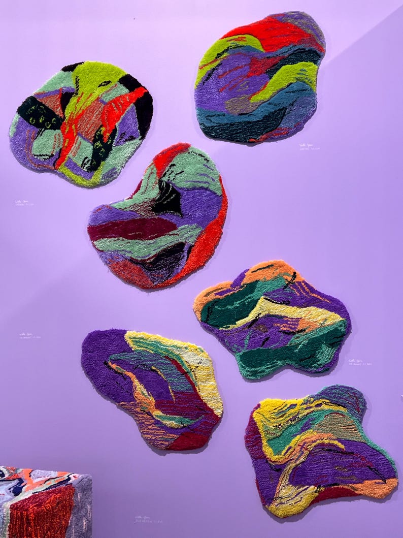 May include: Seven abstract, organic-shaped rugs in various colors, including purple, green, red, and yellow, are displayed against a lavender wall. The rugs have a textured, woven appearance, with each piece showcasing a unique color palette and design.
