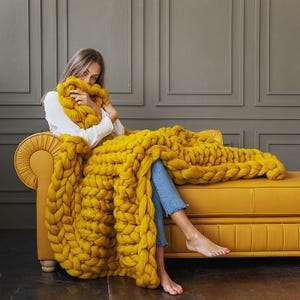 Pode incluir: Uma mulher está sentada em um sofá amarelo, envolvida em um grande cobertor de tricô grosso de cor mostarda. O cobertor é feito de fio grosso e macio e tem uma sensação relaxante e aconchegante.