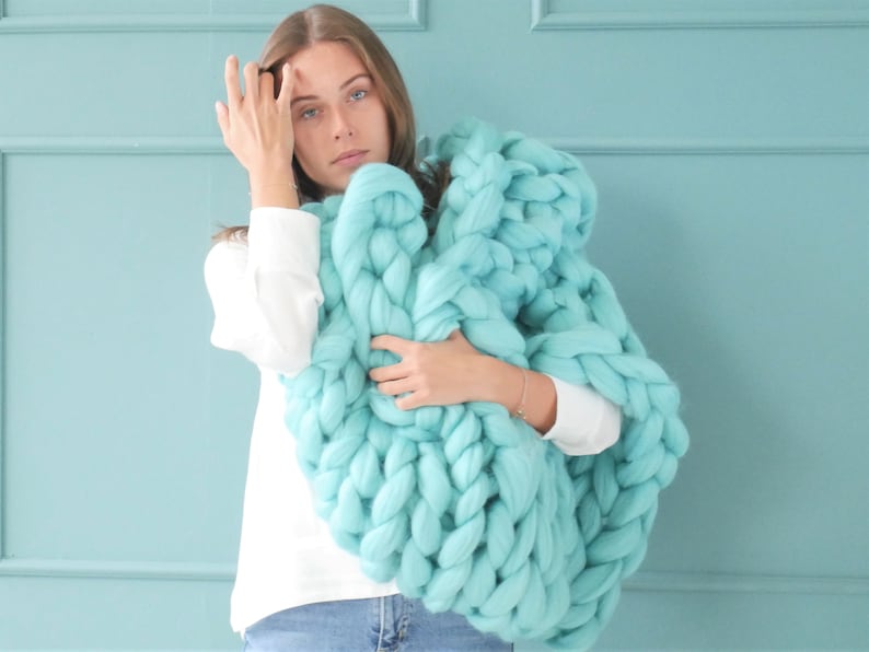 May include: A large, chunky-knit blanket in a soft, light turquoise color. The blanket is made of thick yarn and has a textured appearance. The oversized throw is held by a person wearing a white shirt.