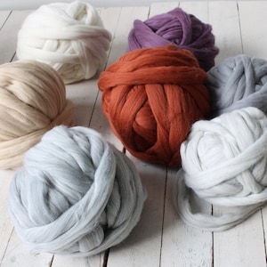Op de afbeelding: Zes bollen garen in verschillende kleuren, waaronder wit, beige, grijs, paars en oranje. Het garen is dik en pluche, perfect om te breien of te haken.