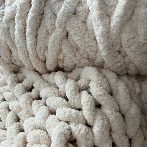 K&ouml;nnte beinhalten: Nahaufnahme einer cremefarbenen, grob gestrickten Decke. Die Decke besteht aus weichem, flauschigem Garn und hat ein strukturiertes, Zopfmuster.