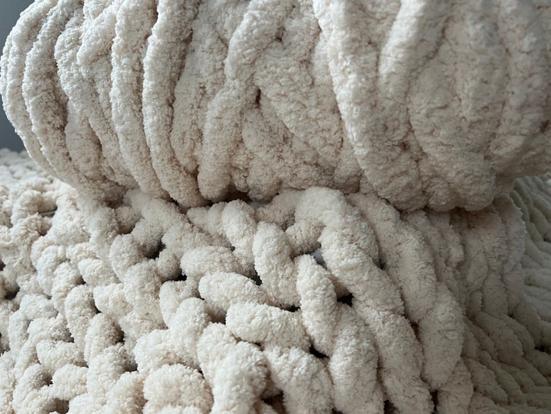 K&ouml;nnte beinhalten: Nahaufnahme einer cremefarbenen, grob gestrickten Decke. Die Decke besteht aus weichem, flauschigem Garn und hat ein strukturiertes, Zopfmuster.