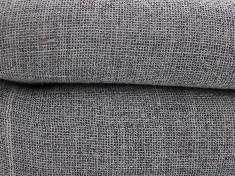 May include: Close-up of a folded, gray linen fabric. The fabric has a coarse, woven texture, with visible threads creating a grid-like pattern. The color is a muted gray, with subtle variations in tone. The fabric appears to be soft and pliable.