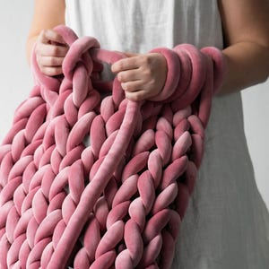 A young woman holding thick, plush velvet yarn, demonstrating the arm knitting technique to create a handmade chunky knit blanket. The cozy, oversized stitches of the giant knit throw are visible as she carefully loops the soft yarn, crafting a warm and stylish home décor piece.