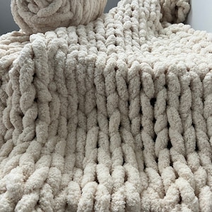 K&ouml;nnte beinhalten: Eine cremefarbene, grob gestrickte Decke mit einer gro&szlig;en, flauschigen Textur. Die Decke ist in der H&auml;lfte gefaltet und eine passende Garnrolle liegt obenauf.
