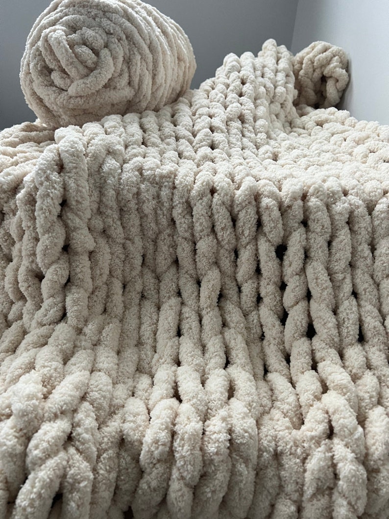 K&ouml;nnte beinhalten: Eine cremefarbene, grob gestrickte Decke mit einer gro&szlig;en, flauschigen Textur. Die Decke ist in der H&auml;lfte gefaltet und eine passende Garnrolle liegt obenauf.
