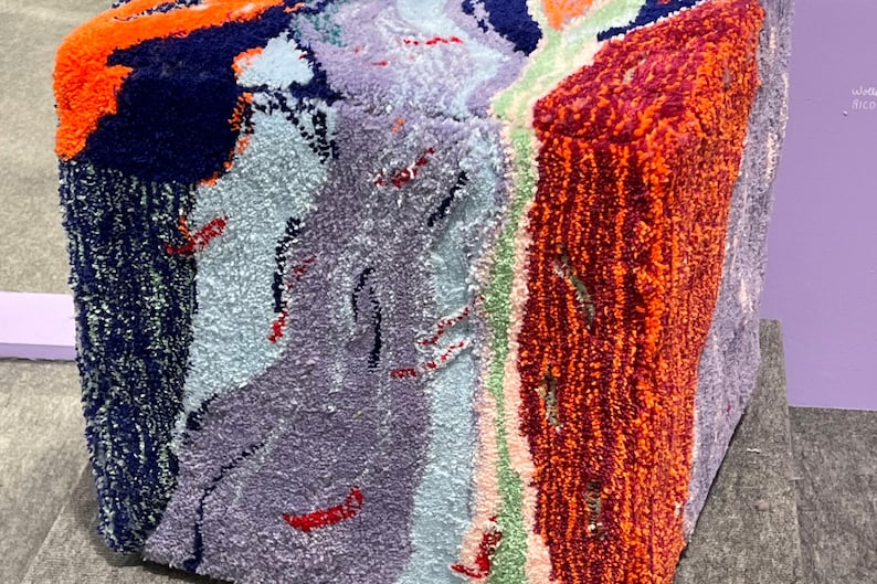 May include: A colorful, textured cube with a variety of colors including orange, navy blue, light blue, purple, and red. The cube appears to be made of a soft, plush material, possibly fabric or yarn.