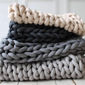 Peut inclure: Une pile de quatre couvertures en tricot &eacute;pais dans des tons de gris et de blanc. Les couvertures sont faites de fil &eacute;pais et doux et ont un aspect confortable et textur&eacute;.