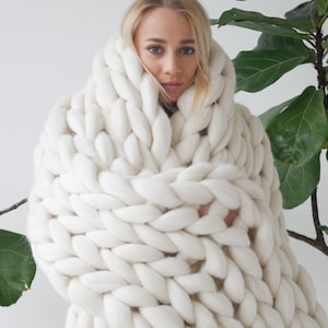 Peut inclure: Une couverture en tricot épais de couleur blanche est drapé sur les épaules d'une personne. La couverture est faite de fil épais et doux et a une sensation confortable et luxueuse.