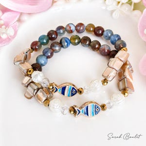 Könnte beinhalten: Zwei Armbänder mit Perlen. Eines hat mehrfarbige runde Perlen. Das andere hat quadratische Muschelkernperlen, weiße Rosenperlen und blau-weiße Fischanhänger. Goldfarbene Akzente sind vorhanden.