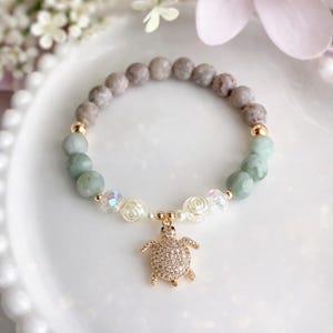 May include: A beaded bracelet featuring a gold-colored turtle charm. The bracelet includes gray and light green beads, with gold accents and small white rose-shaped beads. The turtle charm is encrusted with small, clear stones.