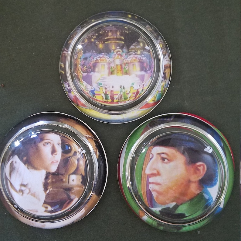 May include: Three glass paperweights with silver rims. The first paperweight shows a colorful scene of a winter wonderland with a building in the center. The second paperweight shows a woman with dark hair looking to the right. The third paperweight shows a man with dark hair and a blue hat looking to the left.