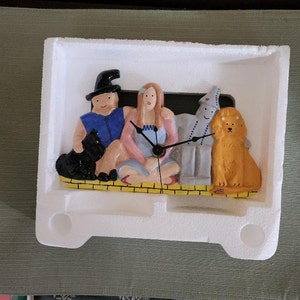 May include: A ceramic clock featuring characters from The Wizard of Oz. The clock depicts the Scarecrow, Tin Man, Lion, Dorothy, and the Wizard. The clock is in a white styrofoam package.