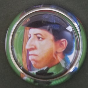 May include: A round, glass paperweight with a silver rim. The paperweight features a portrait of a person wearing a blue hat and a green shirt.