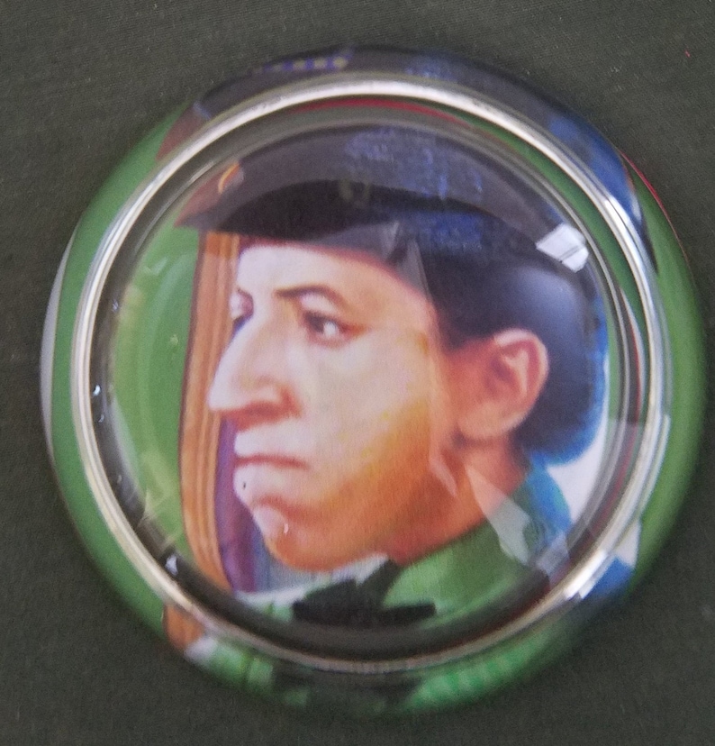 May include: A round, glass paperweight with a silver rim. The paperweight features a portrait of a person wearing a blue hat and a green shirt.