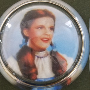 May include: A silver and glass car dashboard ornament featuring a color image of Dorothy from the Wizard of Oz.