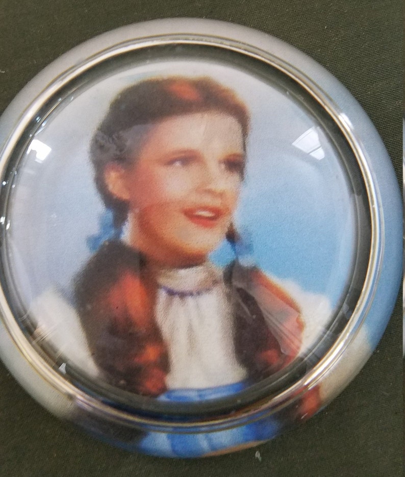 May include: A silver and glass car dashboard ornament featuring a color image of Dorothy from the Wizard of Oz.