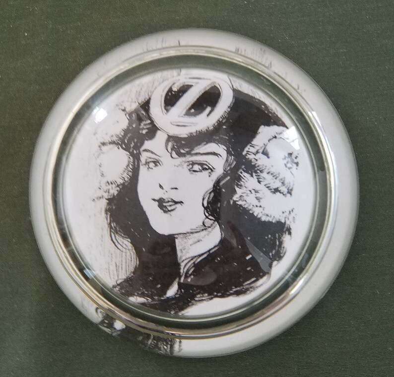 May include: A black and white illustration of a woman wearing a hat with a letter Z on it. The image is inside a round, silver-colored frame.