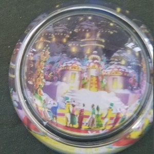May include: A glass snow globe with a winter scene inside. The scene features a small village with snow-covered houses and a group of people gathered in the center. The globe is decorated with a silver rim and a yellow base.