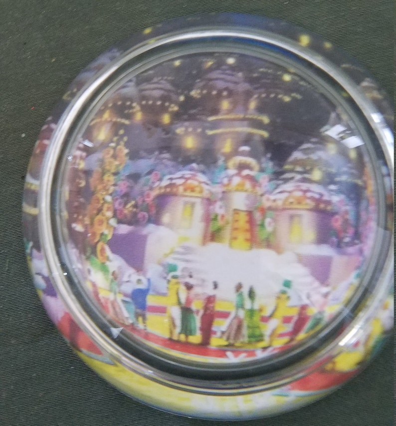 May include: A glass snow globe with a winter scene inside. The scene features a small village with snow-covered houses and a group of people gathered in the center. The globe is decorated with a silver rim and a yellow base.