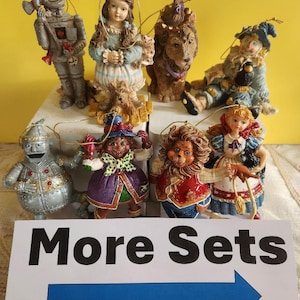 May include: A collection of Wizard of Oz character ornaments, including the Tin Man, Dorothy, the Cowardly Lion, and the Scarecrow. The ornaments are detailed with vibrant colors and are displayed on a white surface. A sign that says "More Sets" is in the foreground.