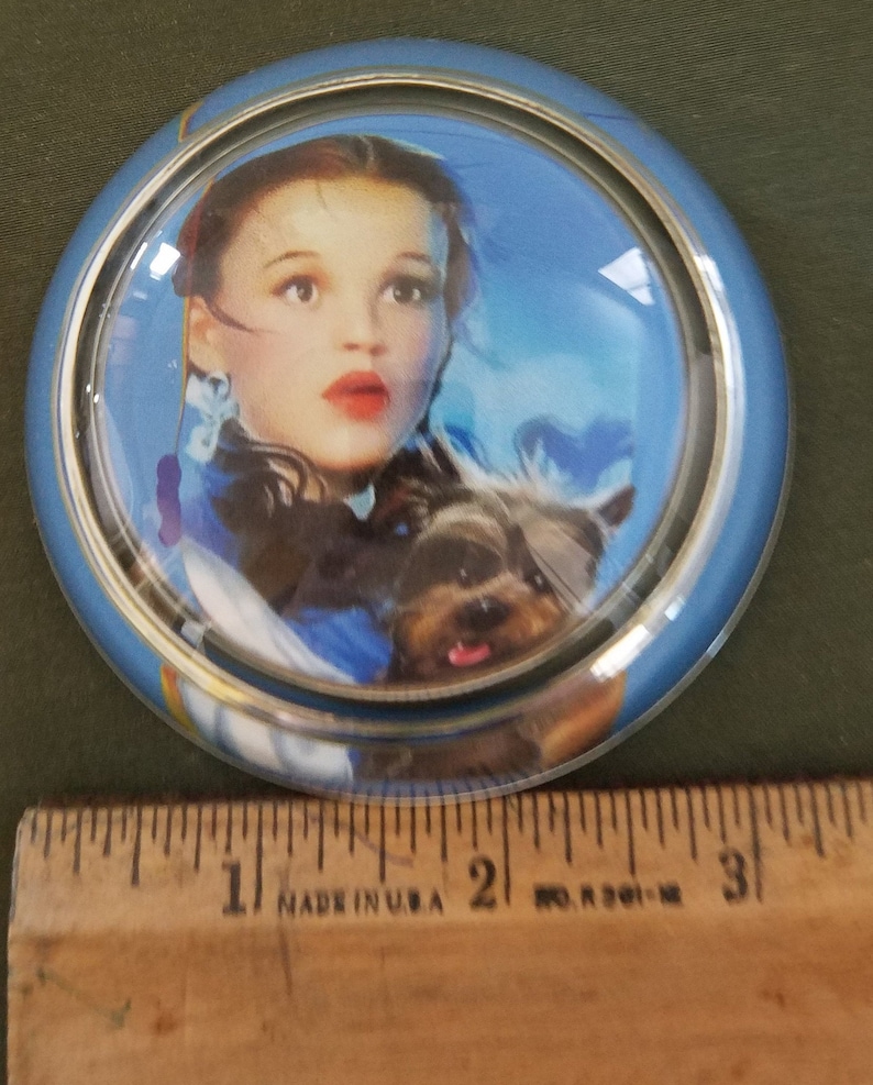 May include: A glass paperweight with a blue background and a picture of Dorothy from the Wizard of Oz holding a small black dog. The paperweight has a silver rim.