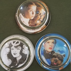 May include: Three round glass paperweights with silver trim. Each paperweight features a different image of Dorothy from the Wizard of Oz. One paperweight shows Dorothy with the Tin Man, another shows Dorothy with Toto, and the last shows a black and white image of Dorothy.