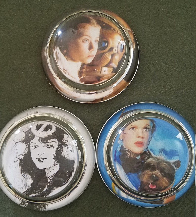 May include: Three round glass paperweights with silver trim. Each paperweight features a different image of Dorothy from the Wizard of Oz. One paperweight shows Dorothy with the Tin Man, another shows Dorothy with Toto, and the last shows a black and white image of Dorothy.