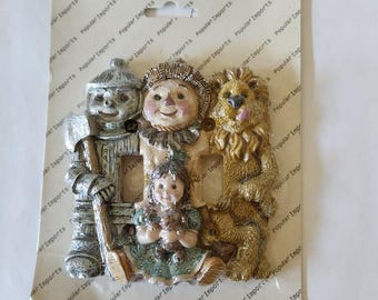 May include: A light switch plate cover featuring a whimsical scene with characters from the Wizard of Oz. The cover features a tin man, scarecrow, lion, and a girl holding a teddy bear. The cover is made of a textured, ceramic-like material and is painted in a variety of colors.