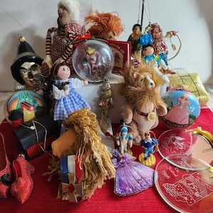 May include: A collection of Wizard of Oz themed ornaments and decorations. Includes a Wicked Witch of the West head, Dorothy doll, ruby slippers, a lion, and a Glinda ornament. Also includes a snow globe and a book.