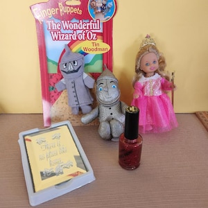 May include: A set of Wizard of Oz themed toys including a finger puppet set featuring the Tin Woodman, a plush Tin Woodman, a doll dressed as Dorothy, and a bottle of red nail polish. The finger puppet packaging features the text "The Wonderful Wizard of Oz". The doll is wearing a pink dress and a gold crown. The nail polish bottle has a black cap and a red glitter finish. The packaging for the finger puppets also includes the text "There is no place like home".