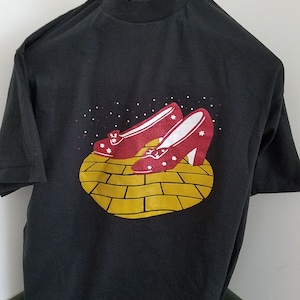 Puede incluir: Camiseta negra con un gráfico de camino de ladrillos amarillos y zapatillas de rubí rojas. El gráfico presenta un par de zapatos rojos con estrellas blancas en un camino de ladrillos amarillos.