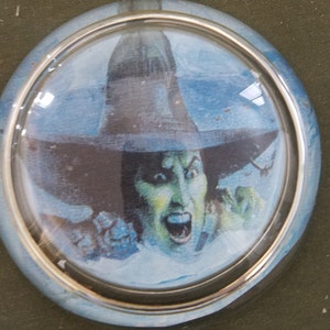May include: A round glass paperweight with a silver rim features a colorful illustration of the Wicked Witch of the West from the Wizard of Oz.