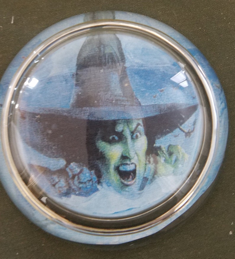 May include: A round glass paperweight with a silver rim features a colorful illustration of the Wicked Witch of the West from the Wizard of Oz.
