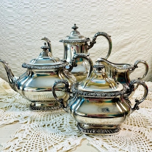 May include: A silver tea set with a teapot, creamer, sugar bowl, and a matching design. The set is on a white crocheted doily.