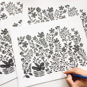 May include: Black and white print of a floral design with birds, bees, and mushrooms. The print is on white paper and is being drawn on with a pencil.