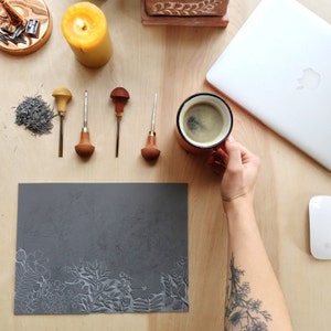 May include: A gray linoleum block with a floral design carved into it. There are four wood carving tools and a cup of coffee on a wooden table.